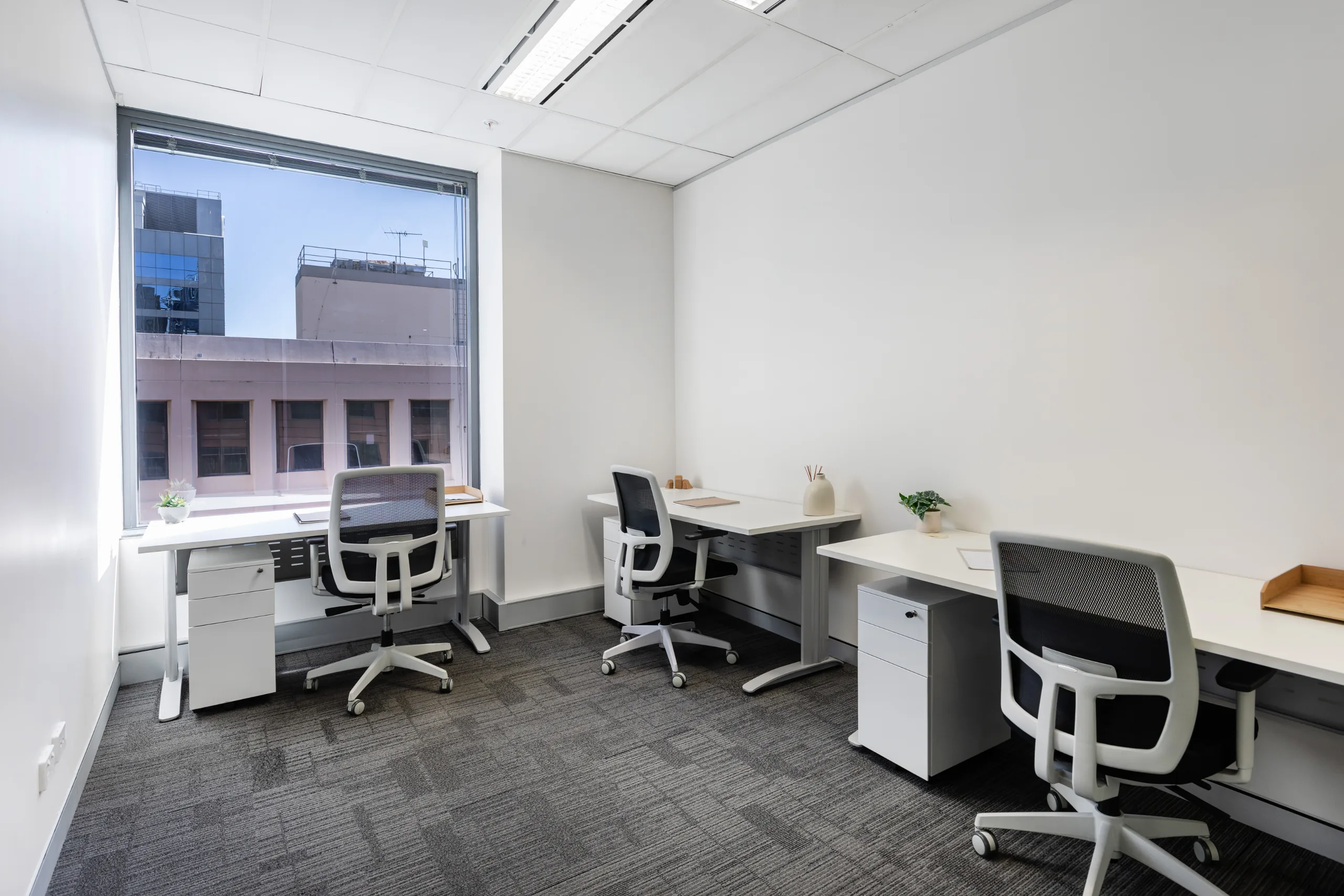 Bright office room featuring three white desks, black ergonomic chairs, and a large window with city views.